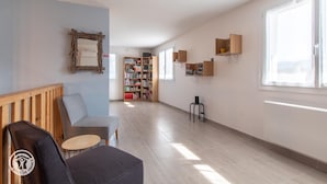 Une pièce spacieuse comprenant un banc en bois, une petite table, une bibliothèque et une fenêtre laissant entrer la lumière naturelle.