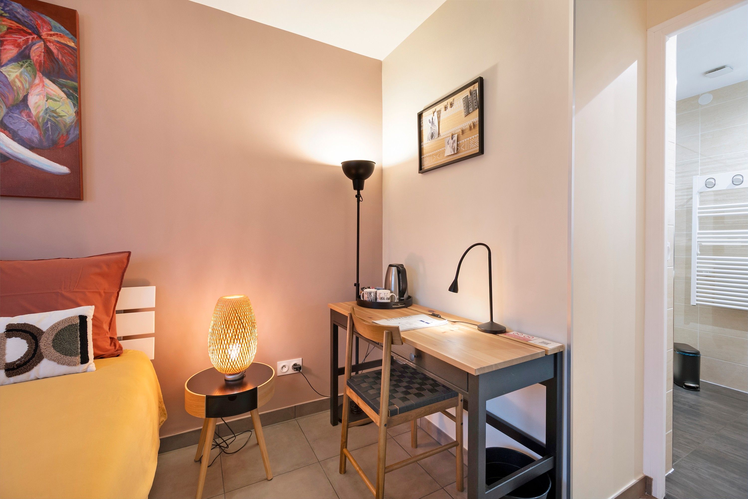 Une petite pièce comprenant un lit, un bureau, une chaise, un lampadaire et un tableau accroché au mur.