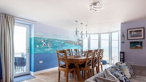 Uma área de refeições com mesa e cadeiras, um quadro de um veleiro na parede e vista para o mar através de grandes janelas.