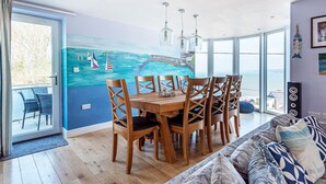 Uma área de refeições com mesa e cadeiras de madeira, um mural com paisagem marítima e grandes janelas com vista para o mar.