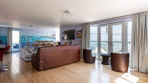 Uma sala de estar ampla com um sofá grande, uma mesa de jantar e cadeiras de vime. É visível uma varanda com vista para o mar através das portas de vidro.