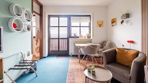 A living room with a sofa, a coffee table, a chair, a dining table, and a window with blinds. There are wall decorations and a radiator. - Apartment 'Bergluft' with Rhine Valley View, Private Terrace and Wi-Fi (Sasbachwalden)