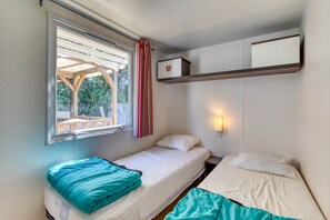 A small room with two beds, a window with a view of an outdoor area, a wall-mounted shelf with a heart-shaped object, and a wall-mounted light.
