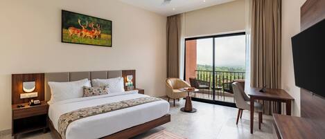 Habitación de hotel moderna con una cama grande, un escritorio, una silla, un televisor, un balcón con vistas y un cuadro en la pared.