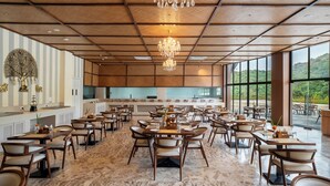 A spacious restaurant with a high ceiling, wooden paneling, and a chandelier. Tables are arranged in a U-shape, facing a large window with a view of greenery.