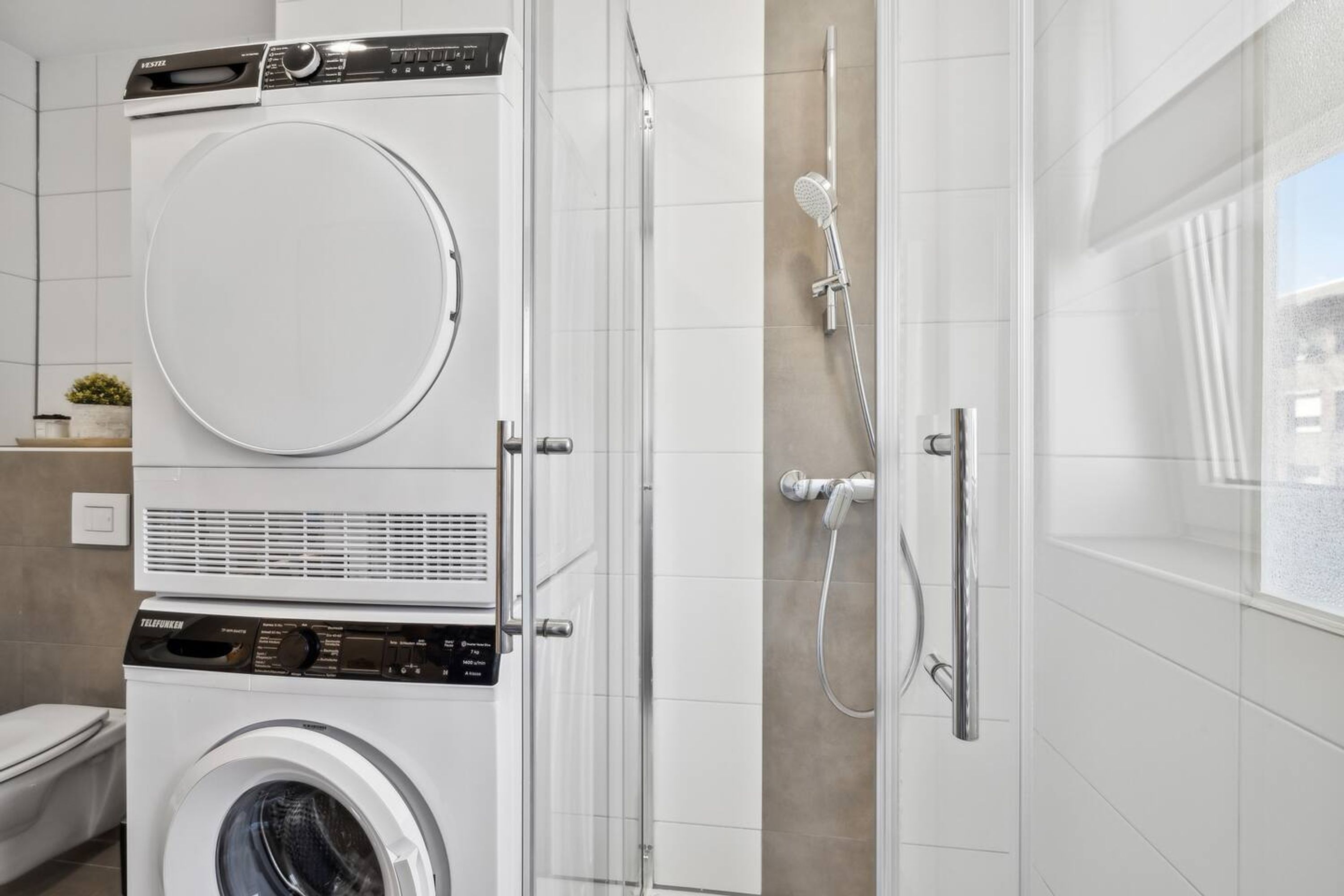 A modern bathroom with a stacked washer and dryer, a glass shower enclosure, and a toilet.