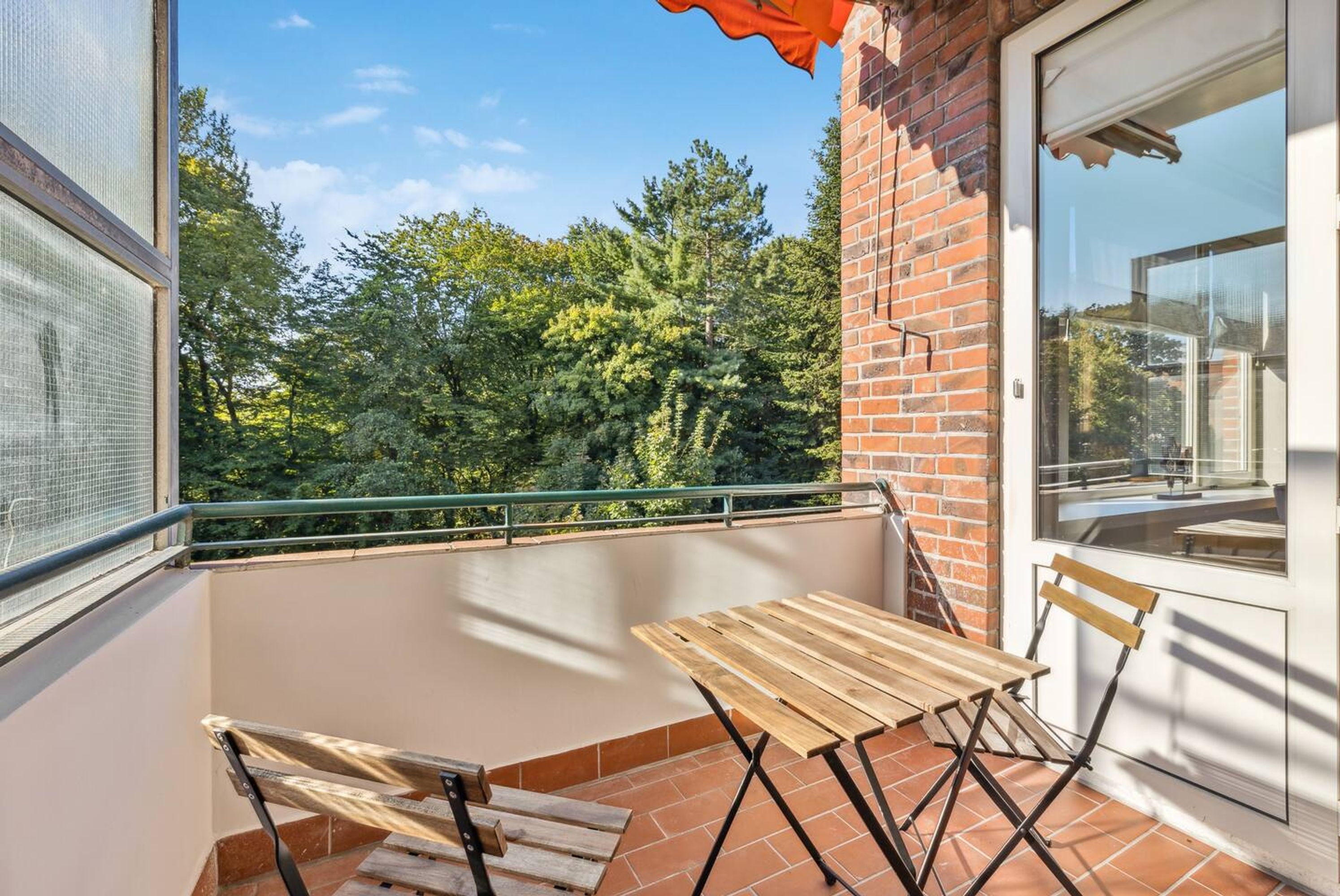 A balcony with a wooden table and chairs, a glass door, and a view of trees.
