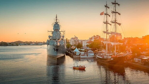 Een groot marineschip ligt aangemeerd in een haven, met een kleinere boot in de buurt.