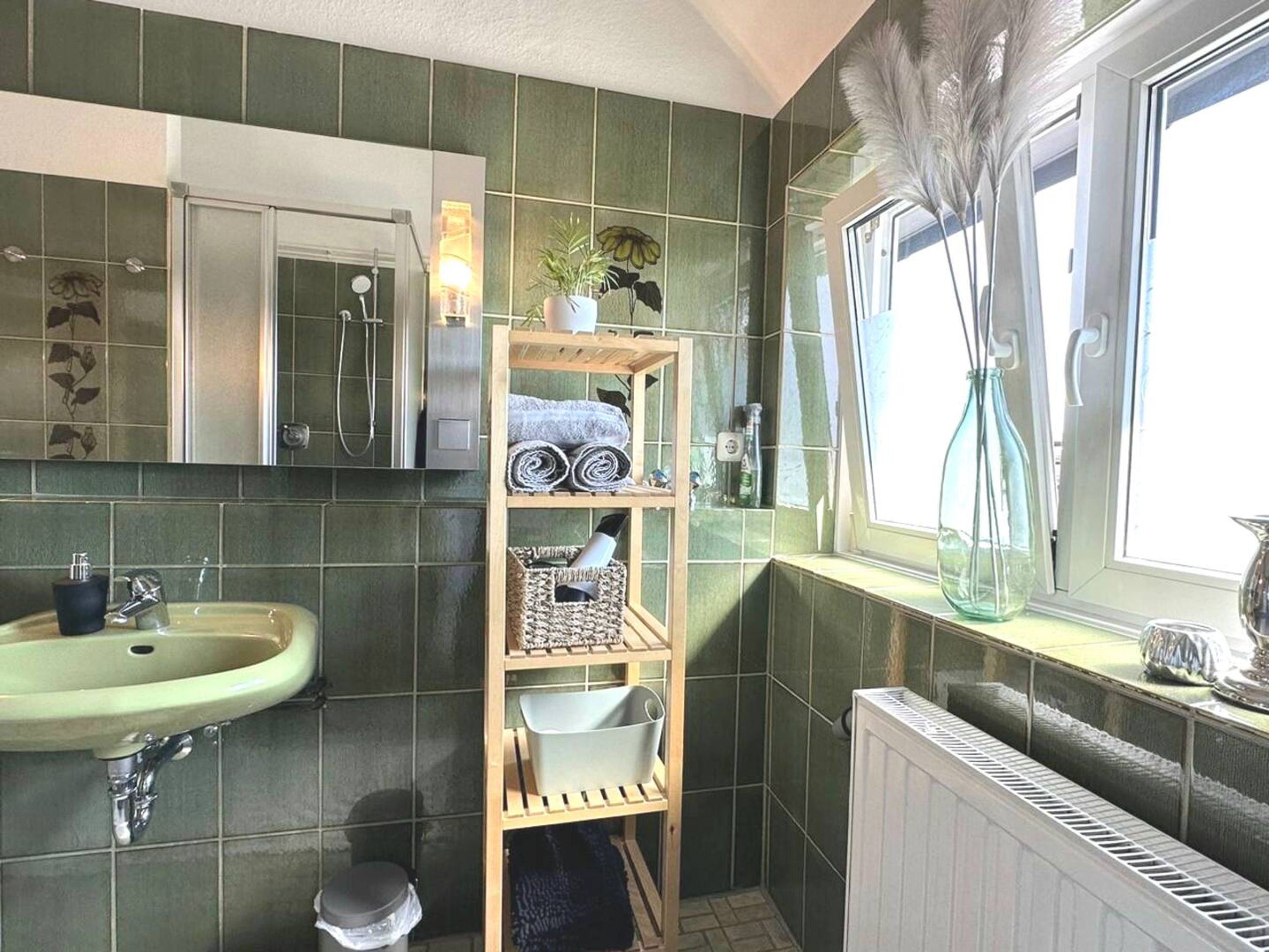 Un baño con pared de azulejos verdes, un estante de madera con toallas, un espejo, un lavamanos y una ventana con vistas.