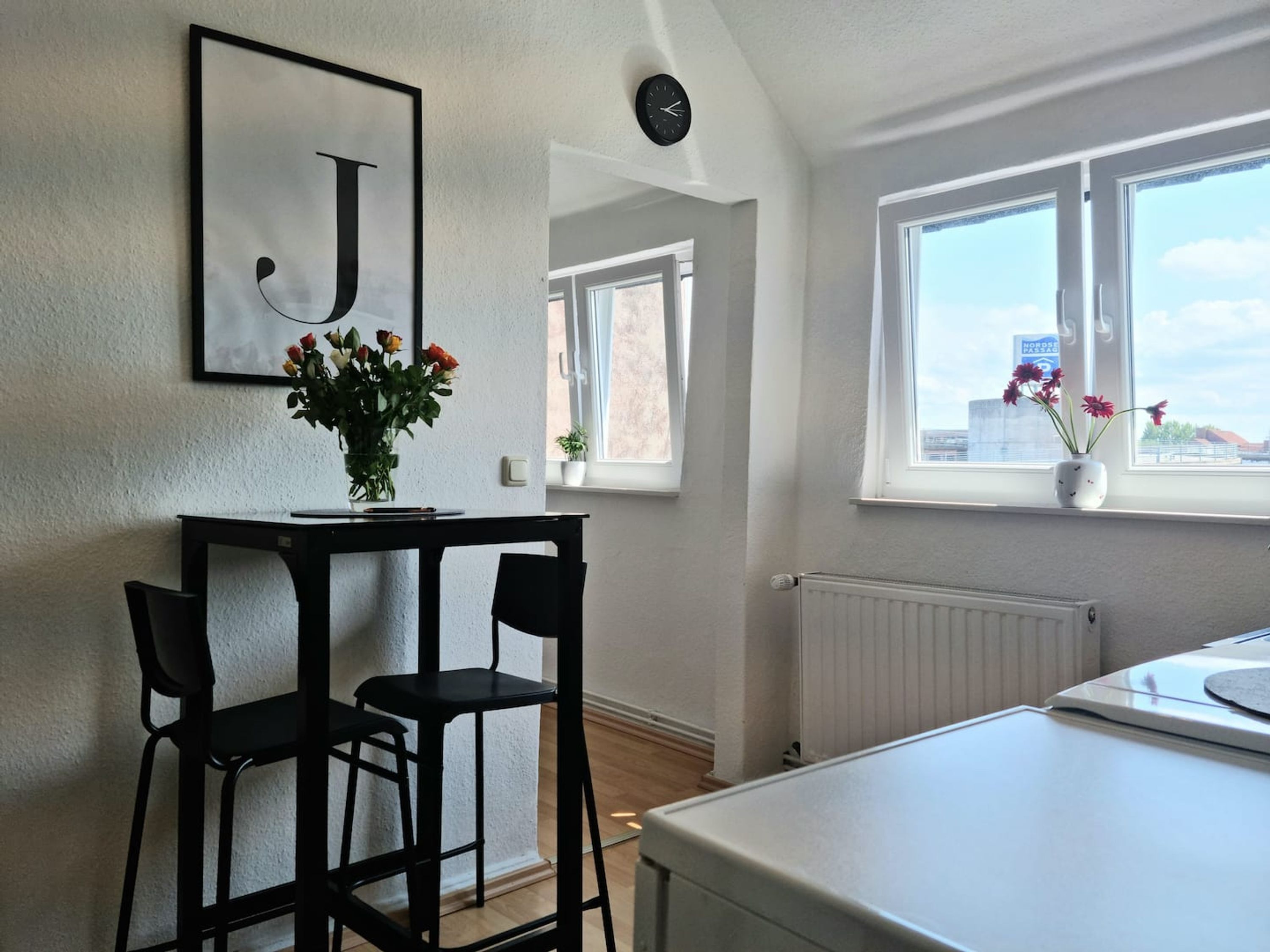 Una cocina moderna con armarios blancos, encimera negra y refrigerador blanco. Hay una ventana con vistas a un paisaje urbano, y un florero con flores en el alféizar.