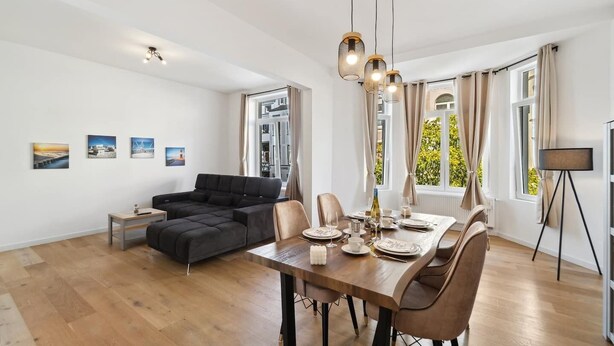 A modern living room with a sofa, dining table, and chairs. There are large windows with curtains, a floor lamp, and artwork on the walls. - Infinity Stay Ferienapartments Rheinstrasse (Wilhelmshaven)