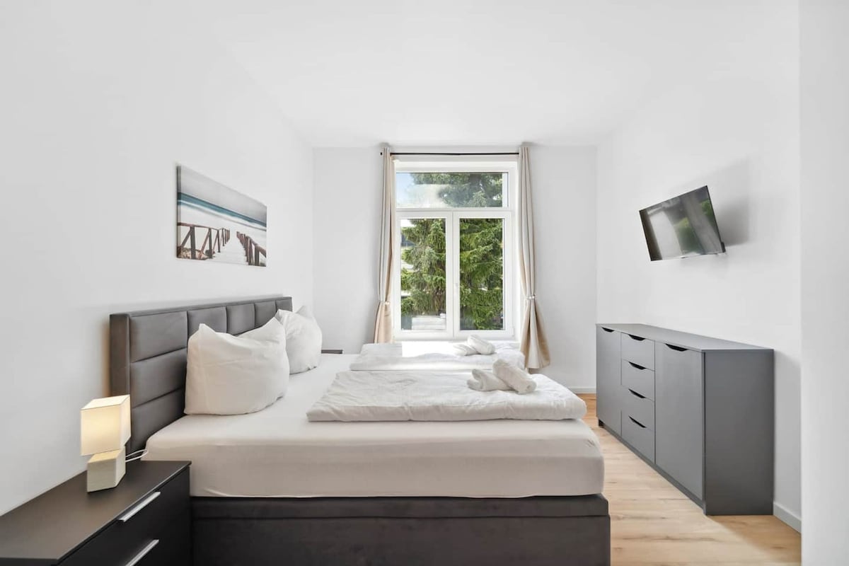 A modern bedroom with a bed, bedside table, TV, and a window with curtains.