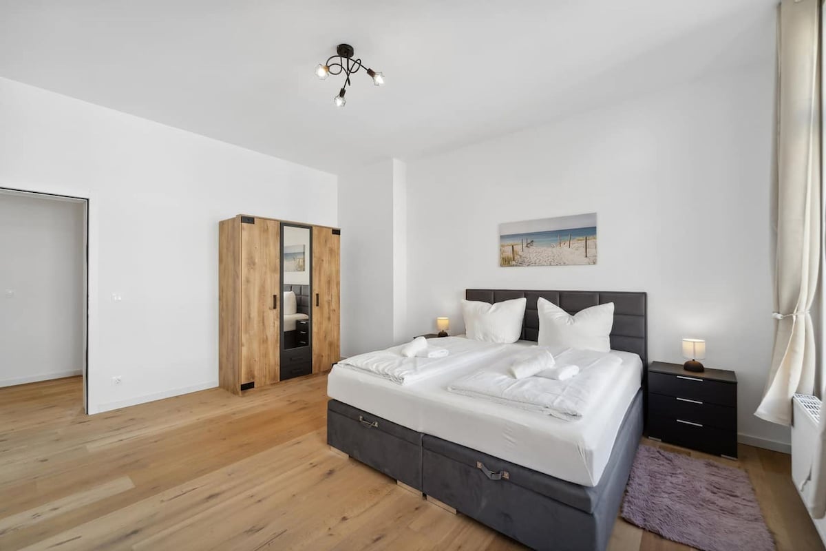 A modern bedroom with a large bed, a wooden wardrobe, a nightstand, and a ceiling light.