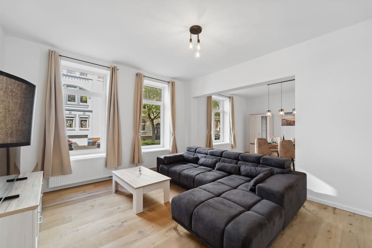 A modern living room with a sectional sofa, a coffee table, and a TV. Large windows with curtains provide natural light and views of the outside.
