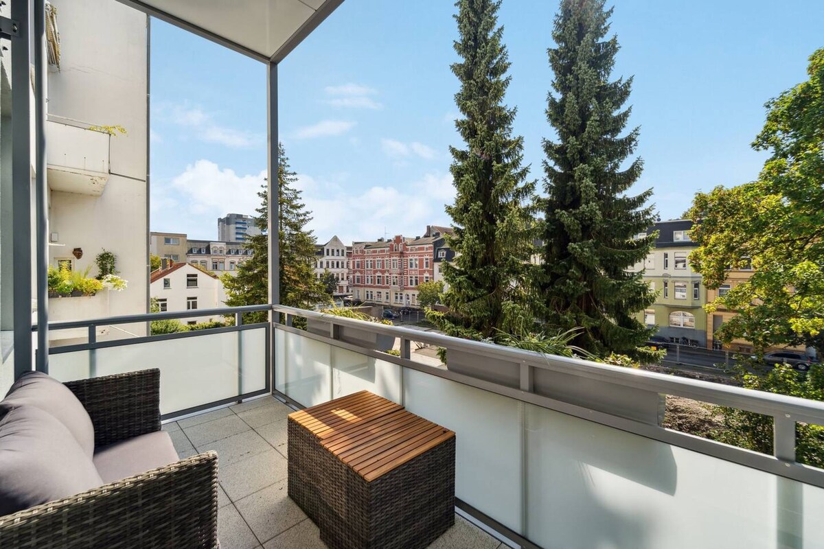 A balcony with wicker furniture, a wooden table, and a view of buildings and trees.