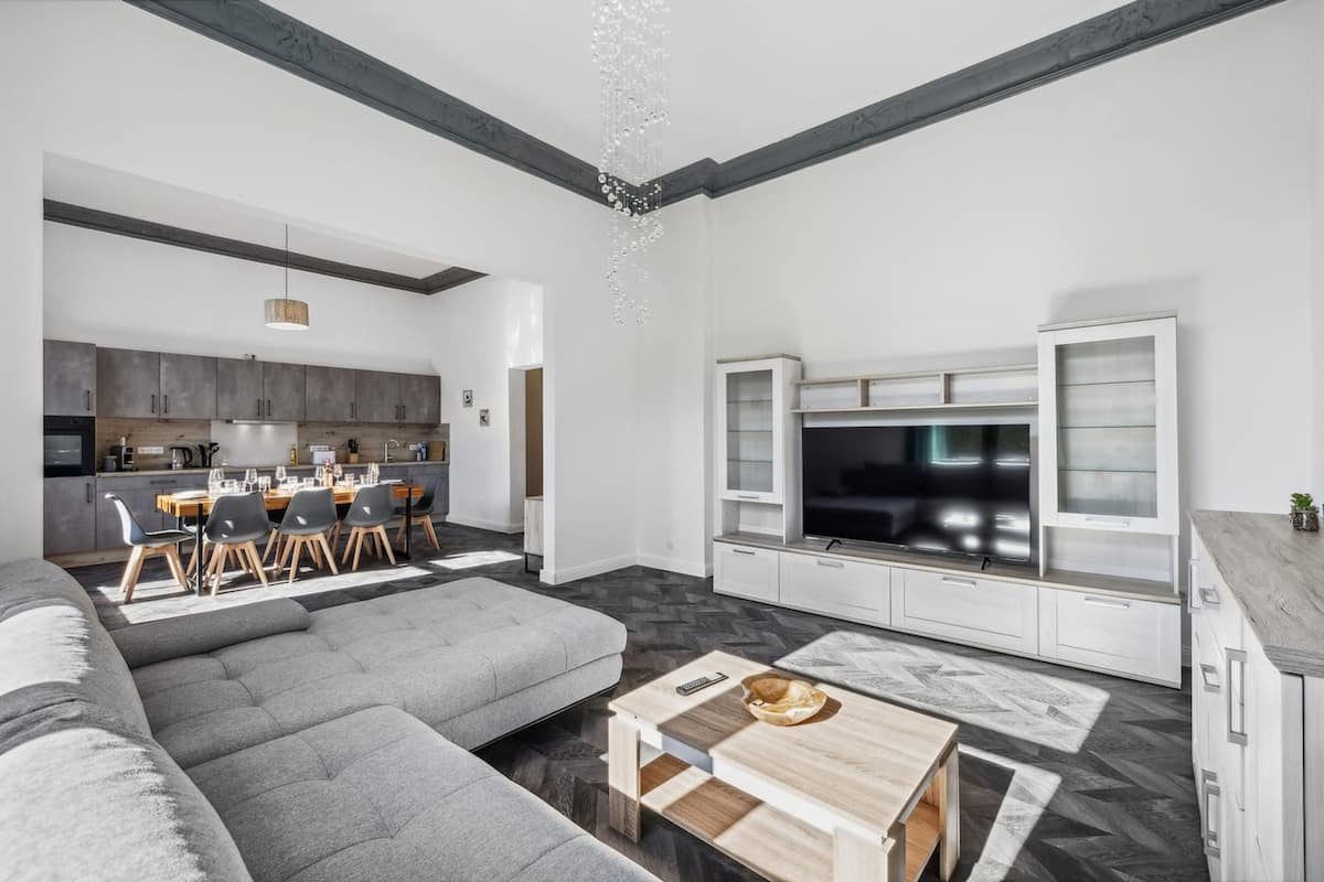 Un salon moderne avec un canapé d’angle gris, une table basse en bois et un téléviseur posé sur un meuble blanc.