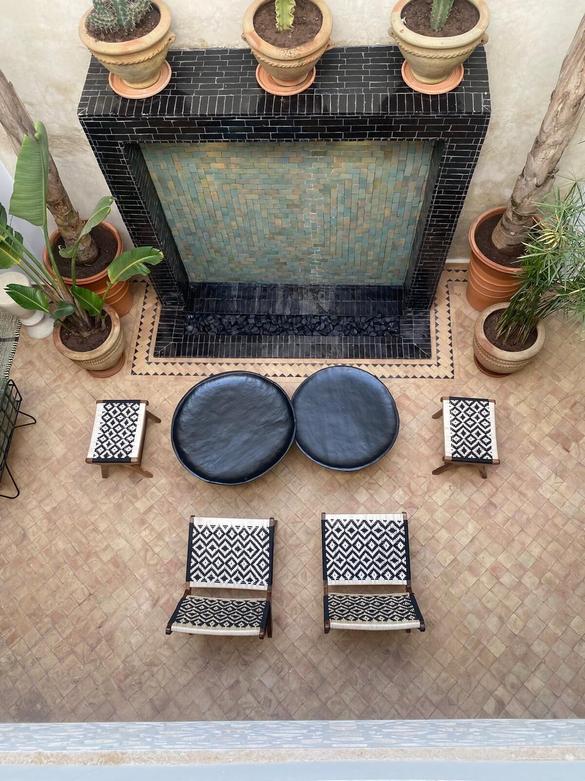 A tiled patio with a black tiled fireplace, surrounded by potted plants and a seating area with patterned cushions.