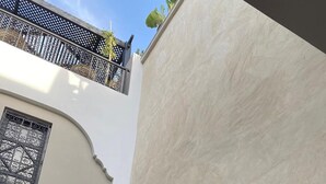 Ein Innenhof mit drei Kakteen in Töpfen vor einer weißen Wand, ein Balkon mit Gittergeländer und ein schwarzer Fliesenbelag am Eingang.