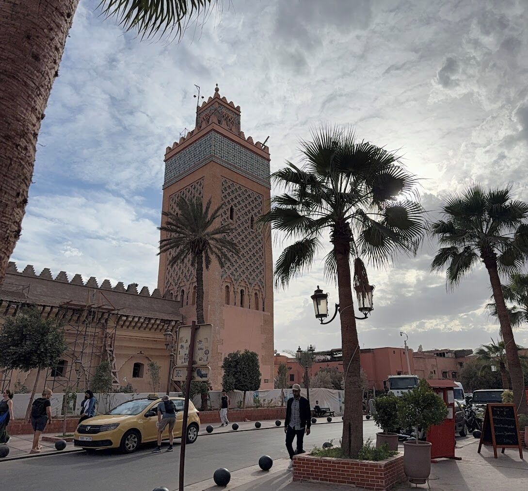 Un edificio histórico con una torre alta, palmeras y una calle con peatones y vehículos.
