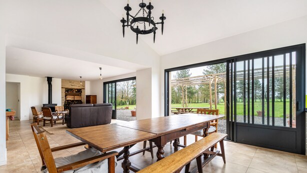 A spacious dining area with a large wooden table and benches, a modern kitchen in the background, and large windows offering a view of the outdoors.