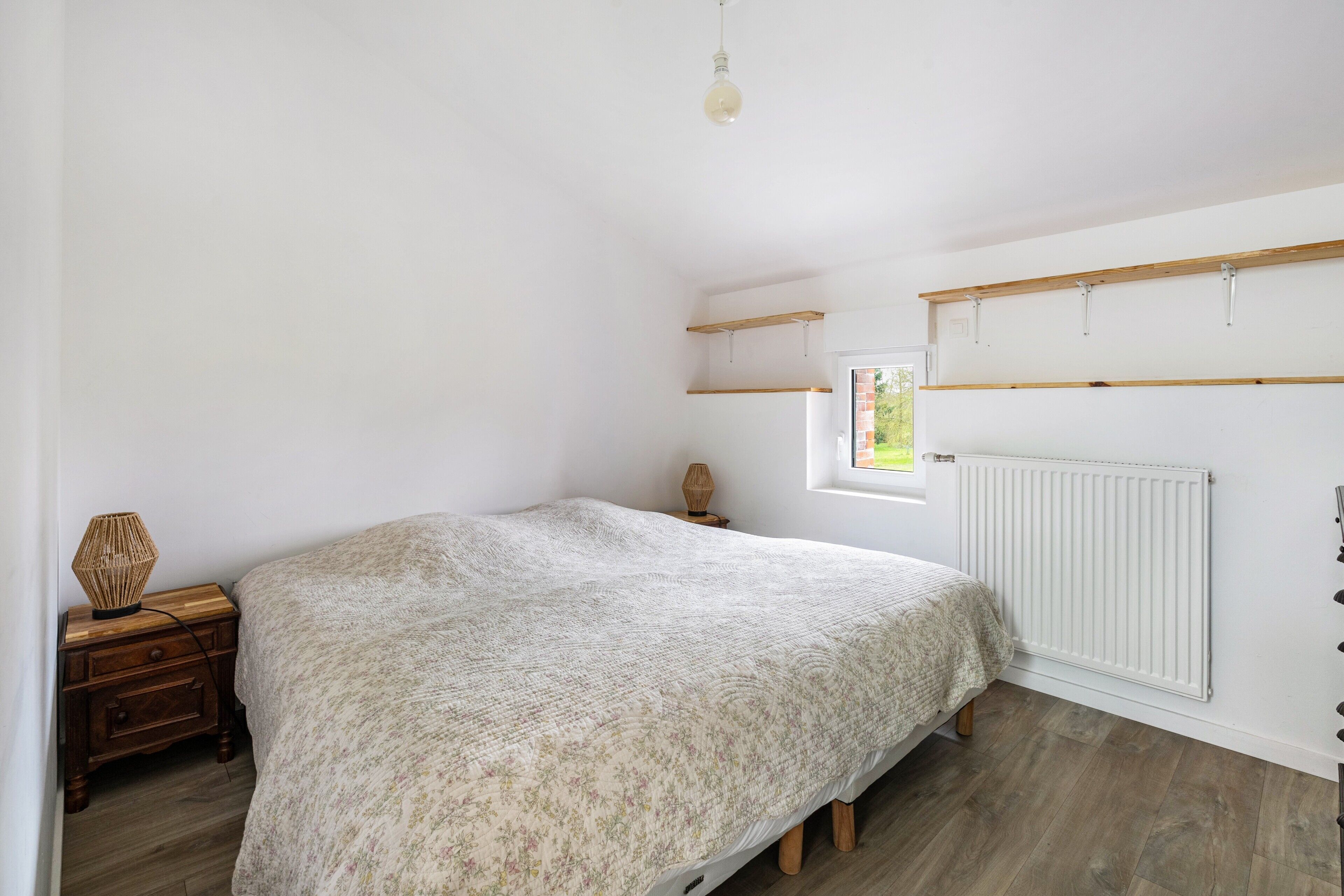 Een slaapkamer met een bed, nachtkastje, lamp, raam en radiator.