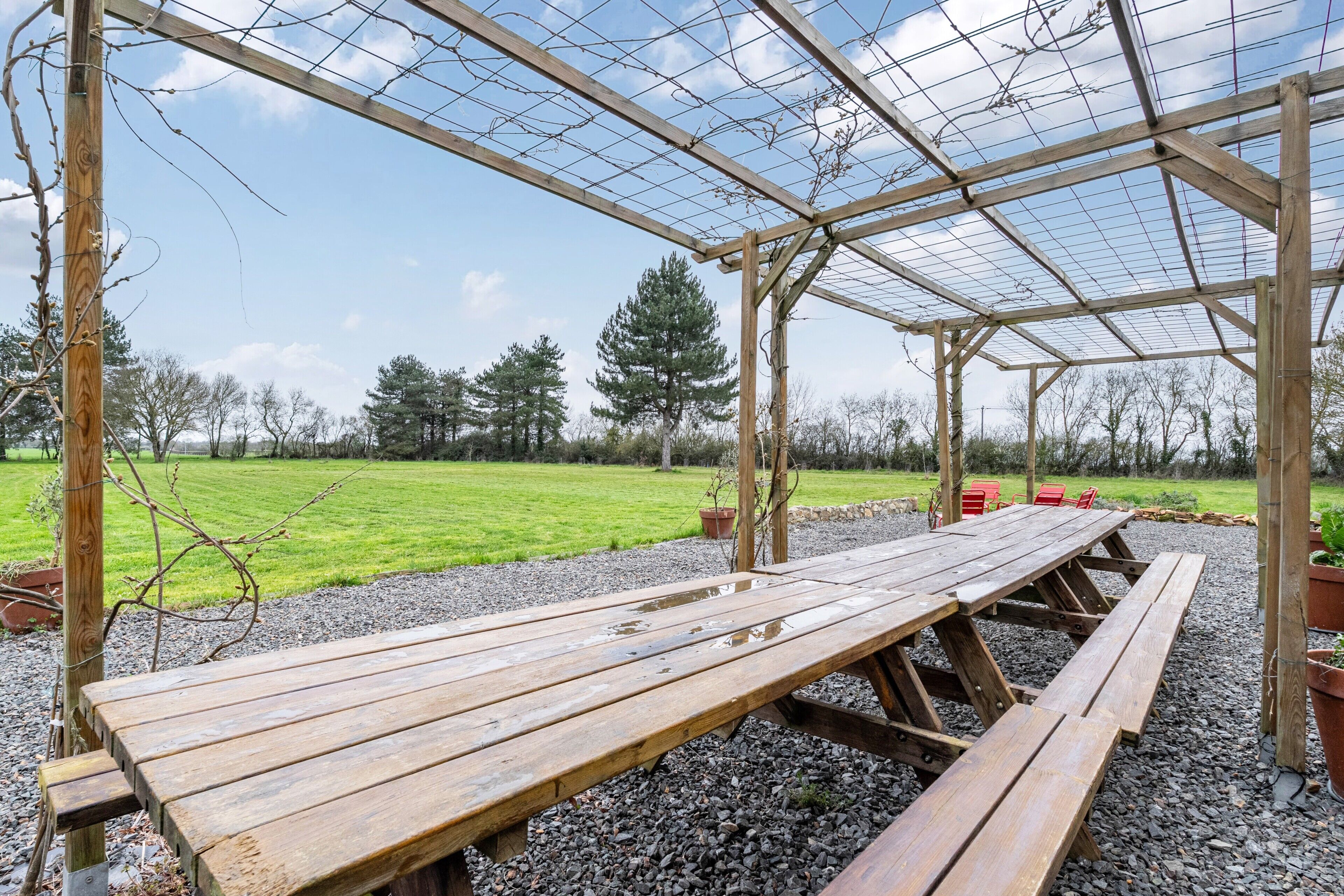 Una pergola in legno con tavolo da picnic e panche, circondata da ghiaia e un prato.