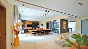 A spacious living area with a large dining table, a sofa, and a coffee table. There is a kitchen area in the background with a TV and cabinets. - Villa Majestueuse - Luxury, Heated Pool & Jacuzzi in Marrakech (Marrakech)