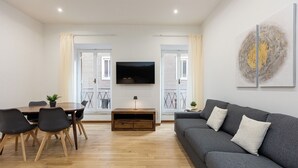 Un soggiorno moderno con un divano grigio, un tavolo da pranzo in legno e sedie. Un televisore a schermo piatto è montato a parete e c'è un piccolo tavolino con una lampada.