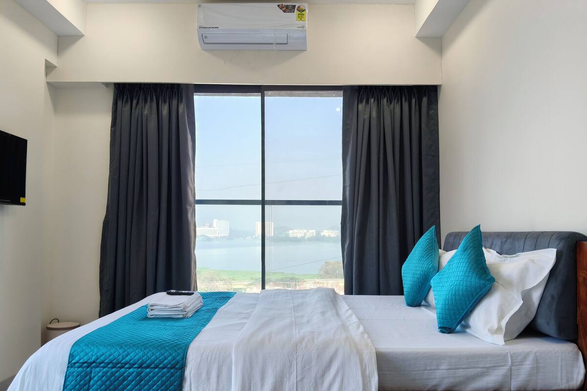 A hotel room with a bed, a view of a body of water, and a mounted air conditioner.