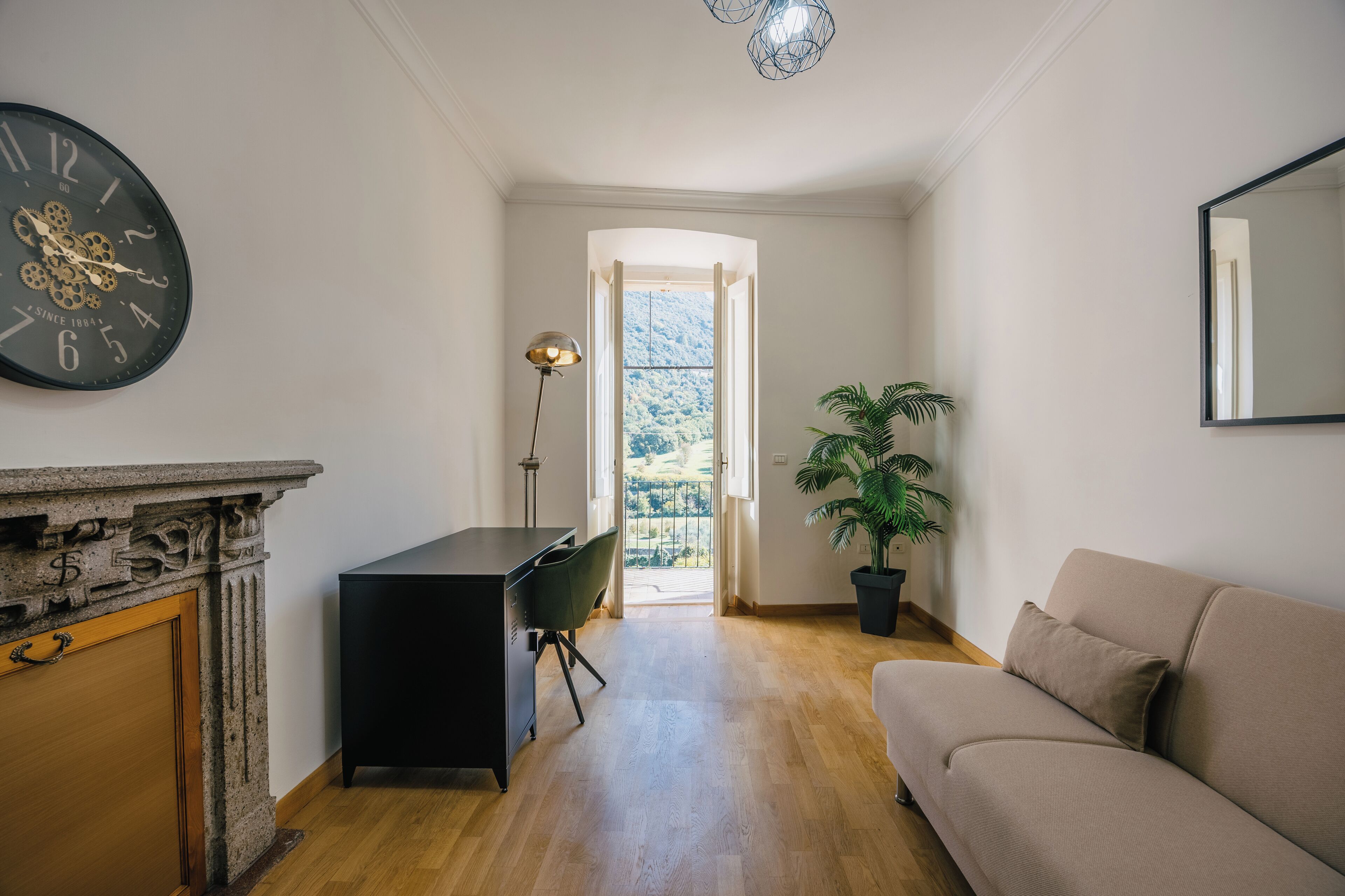 A room with a wooden floor, a black desk, a beige sofa, a potted plant, a wall clock, and a window with a view of greenery.
