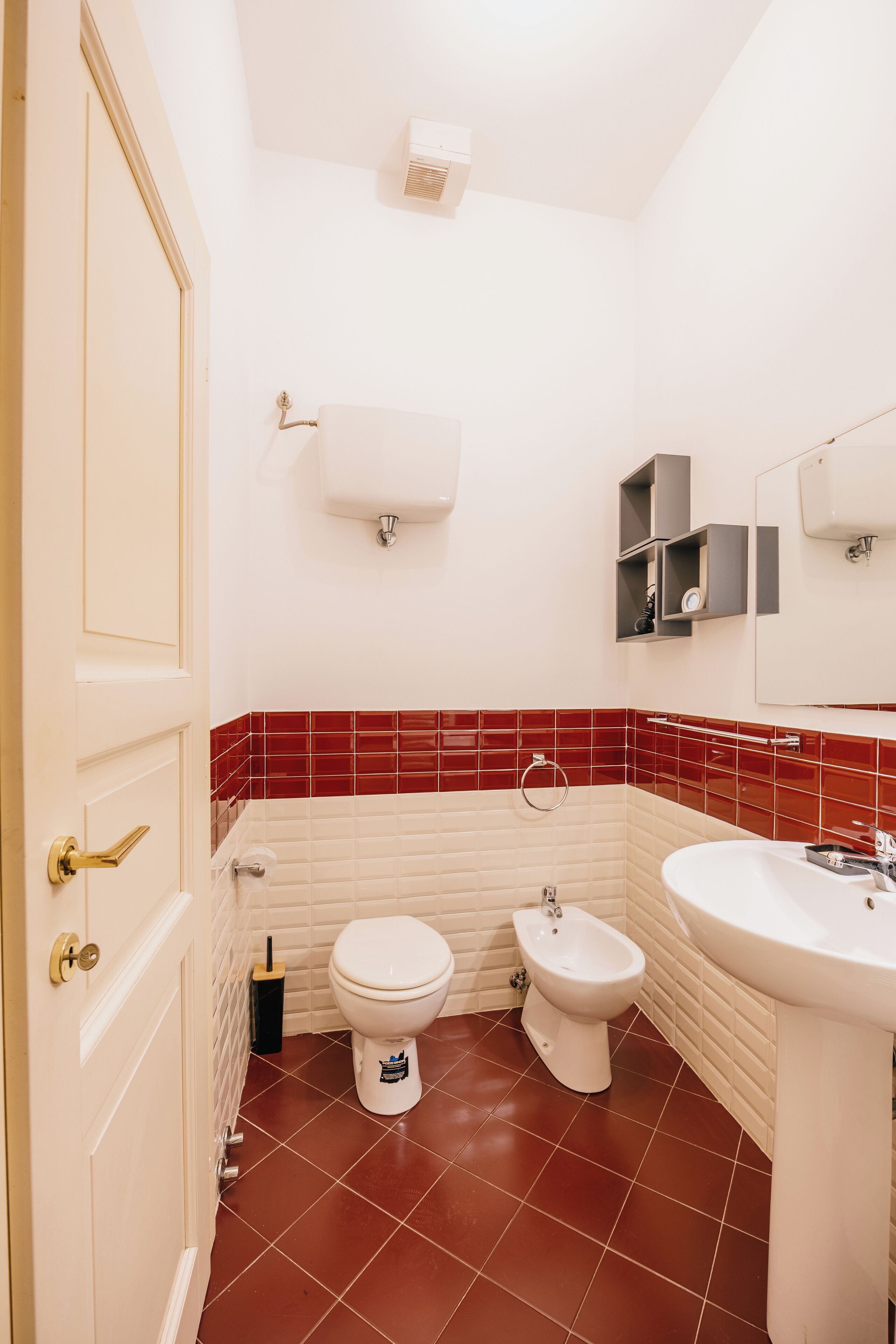 Un baño con inodoro, bidé y lavamanos, con paredes de baldosas rojas y una puerta blanca.