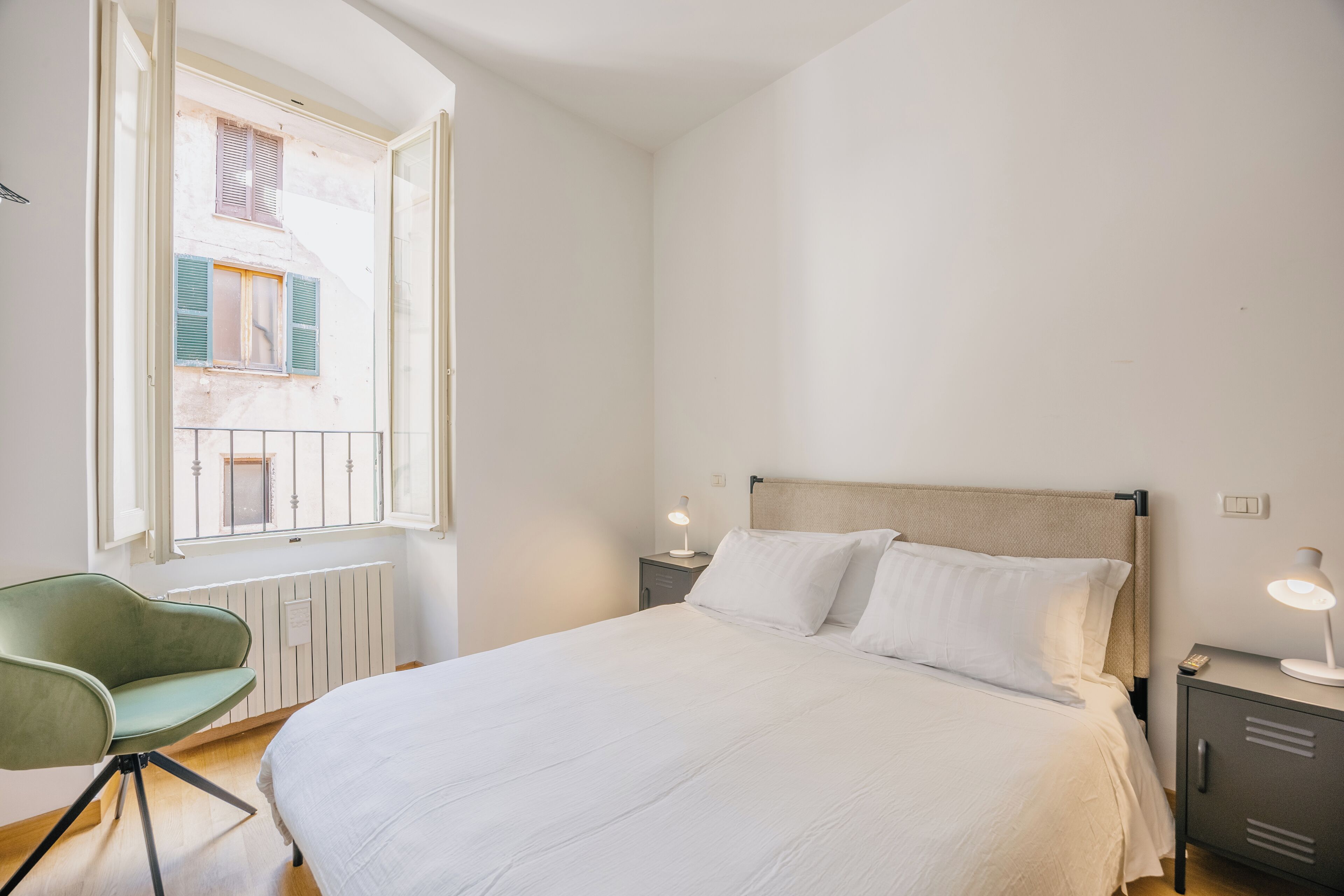 Ein Schlafzimmer mit Bett, Nachttischlampen, einem Sessel, einem Heizkörper und einem Fenster mit Fensterläden.