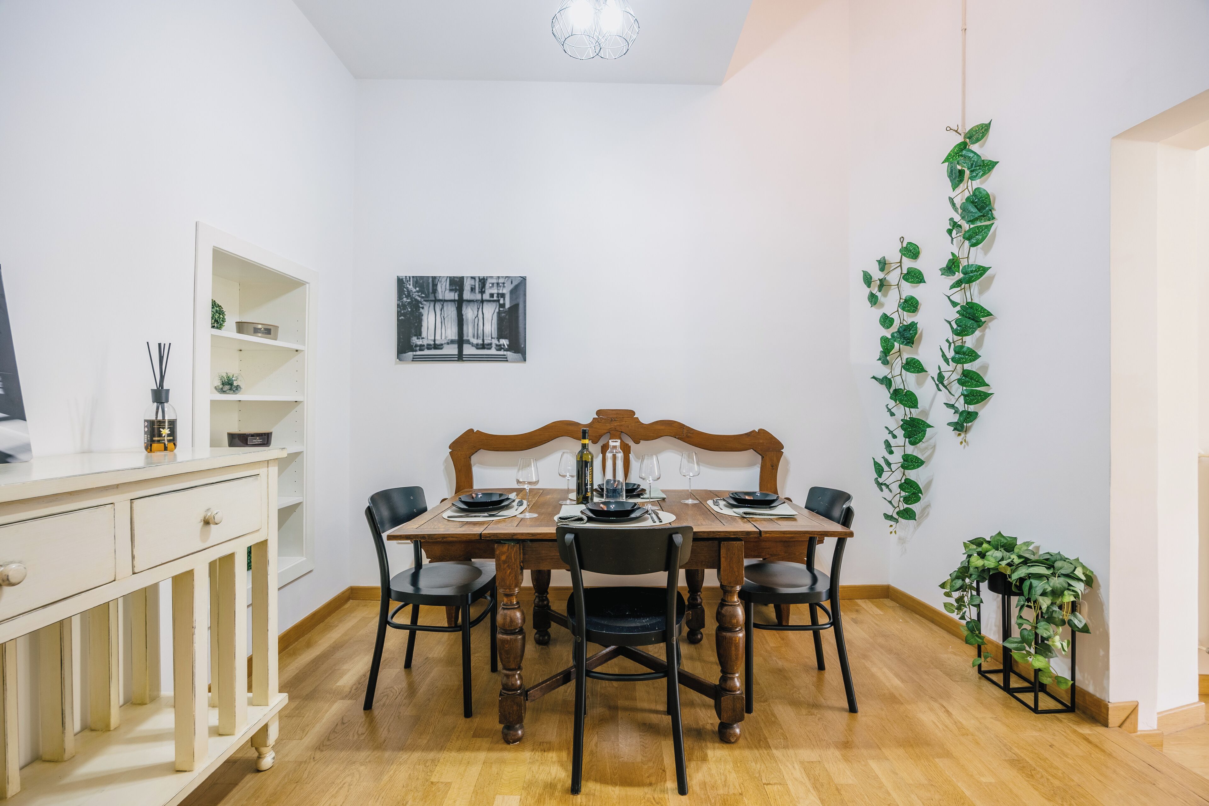 Zona pranzo con tavolo in legno apparecchiato per quattro, circondato da sedie nere. A sinistra, un armadietto bianco con cassetti e, sopra il tavolo, una mensola a parete con oggetti decorativi.