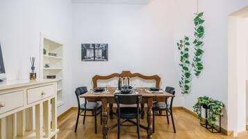 Zona pranzo con tavolo in legno apparecchiato per quattro, circondato da sedie nere. A sinistra, un armadietto bianco con cassetti e, sopra il tavolo, una mensola a parete con oggetti decorativi.