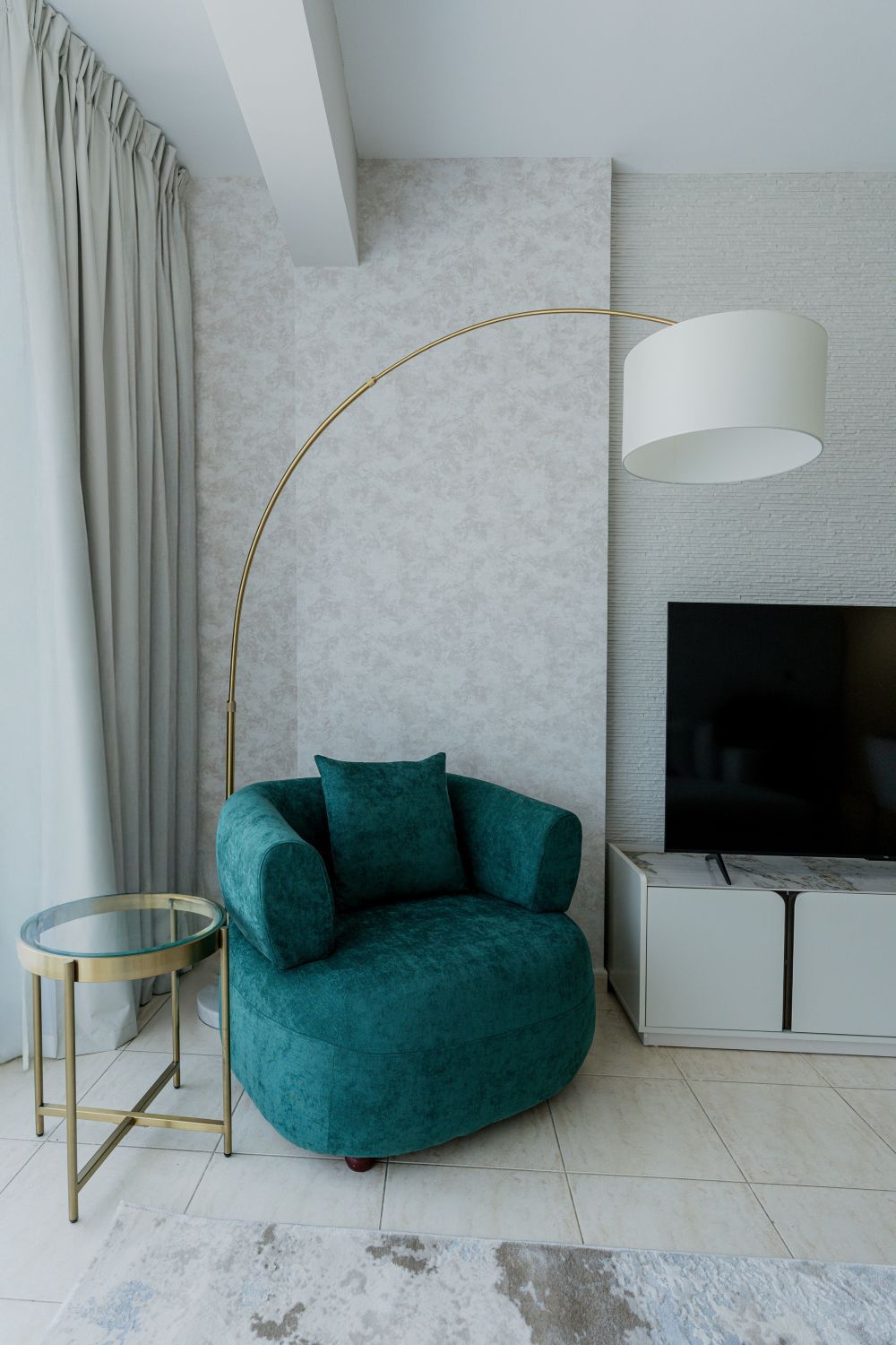 A teal armchair with a gold floor lamp, a TV on a white cabinet, and a glass side table.