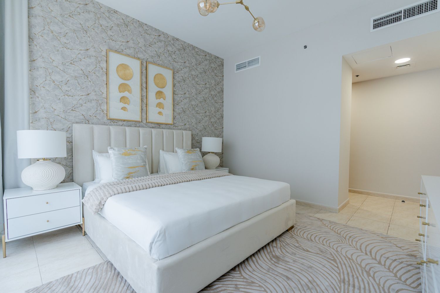 A modern bedroom with a marble wall, a large bed, bedside tables with lamps, and a patterned rug.