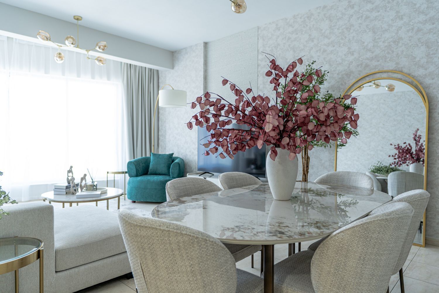 Un salon moderne avec une table ronde en marbre, des sièges confortables, un fauteuil turquoise et un grand vase rempli de fleurs rouges.