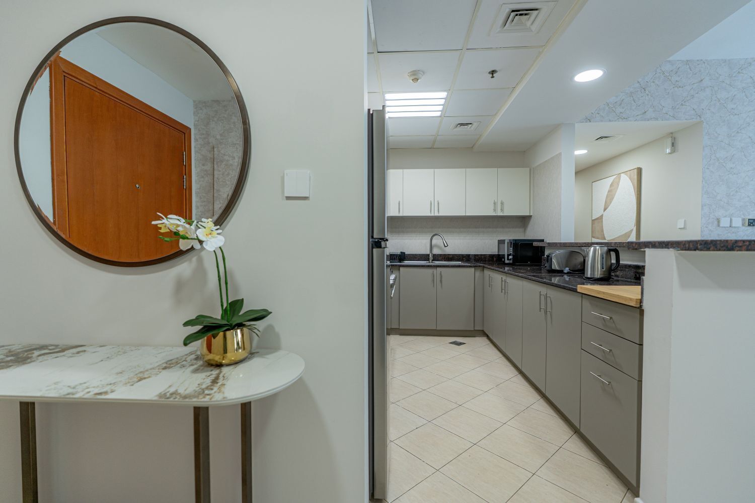 Une cuisine moderne avec des meubles blancs, un plan de travail en marbre et un évier. Un miroir rond est accroché au mur et une plante en pot est posée sur une petite table.