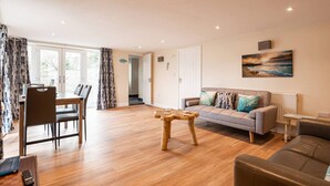 A spacious living room with a wooden floor, a sofa set, a dining area with chairs, and a view through the glass door. - Modern ground floor apartment, near the beach (Ceredigion)