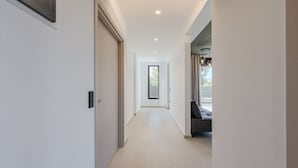 A modern hallway with a neutral color palette, recessed lighting, and a view into a room with a window and a dark-colored sofa. - Villa Allargo - with Private Pool (Gerani)