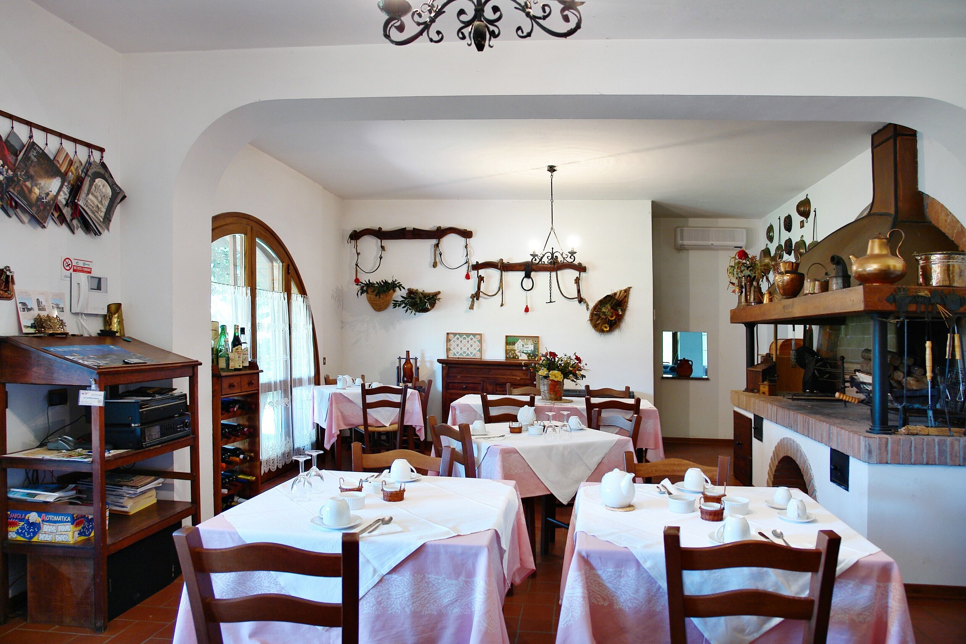 A dining area with tables set for four, wooden chairs, a chandelier, and a kitchen area with a chimney.