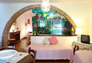Ein Zimmer mit einem Ziegelbogen, einem Bett mit rosa Bettdecke, einem Fernseher auf einem kleinen Tisch und einem großen Fenster mit einer farbenfrohen Wandmalerei.
