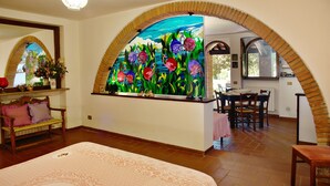 Una habitación con cama, zona de comedor y un amplio ventanal con un cuadro de flores.