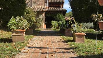 A brick pathway leading to a rustic building with a tiled roof, surrounded by greenery and potted plants.