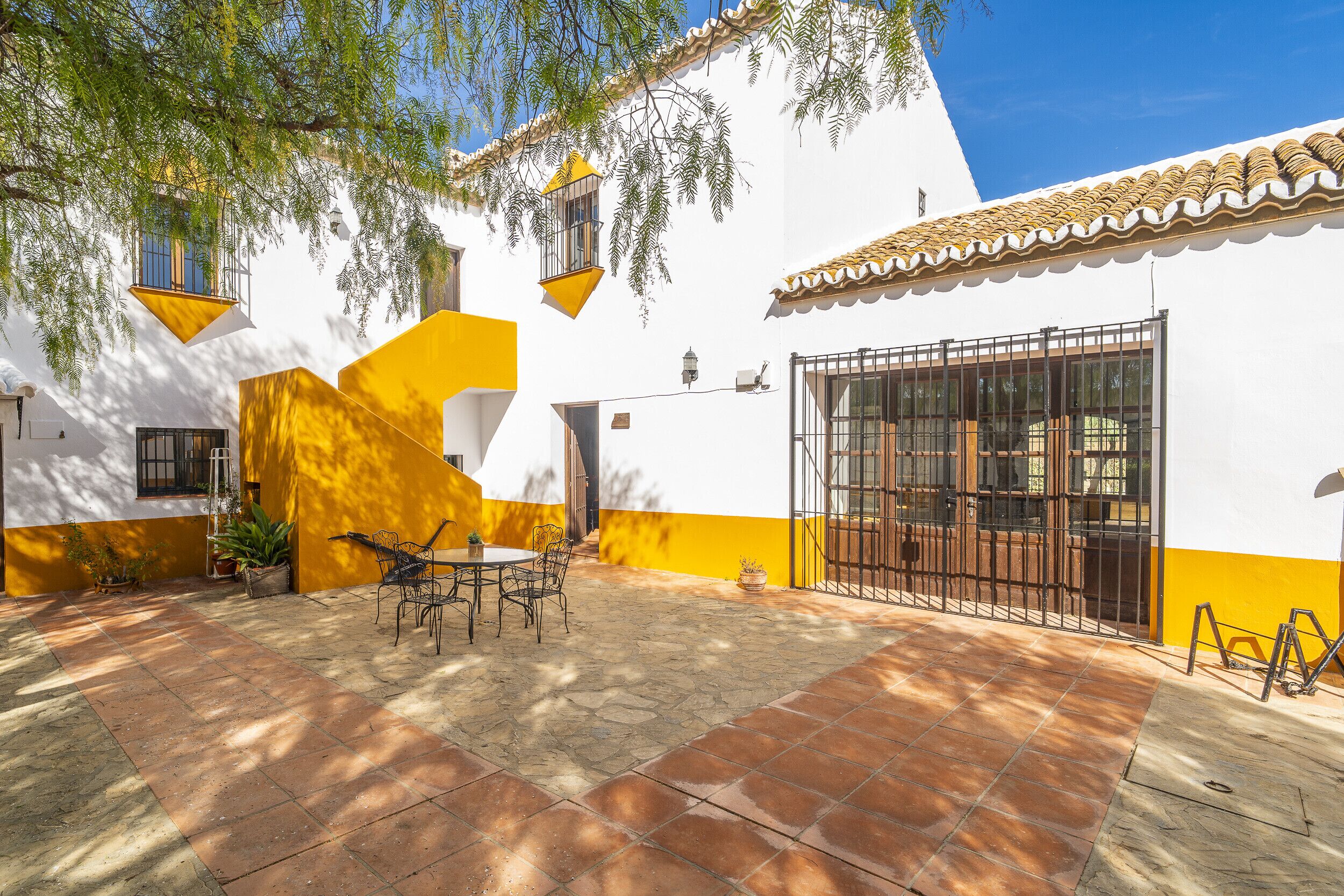 A courtyard with a tiled patio, a table with chairs, and a yellow and white building with a tiled roof.