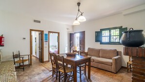 A dining area with a wooden table and chairs, a beige sofa, and a large pot on a stand. - Cubo's Casa Rural San Cristobal Cortijo el Puntal / rural / communal pool / large outdoor areas / views / free wifi (Teba)