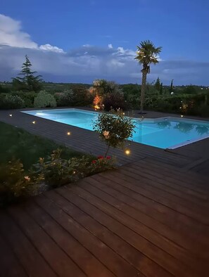 Une piscine rectangulaire entourée d’une terrasse en bois, bordée de différentes plantes et arbres.