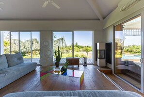 Un salon spacieux avec un canapé gris, une table basse en verre aux couleurs vives, un téléviseur et de grandes fenêtres donnant sur une piscine et un espace verdoyant.