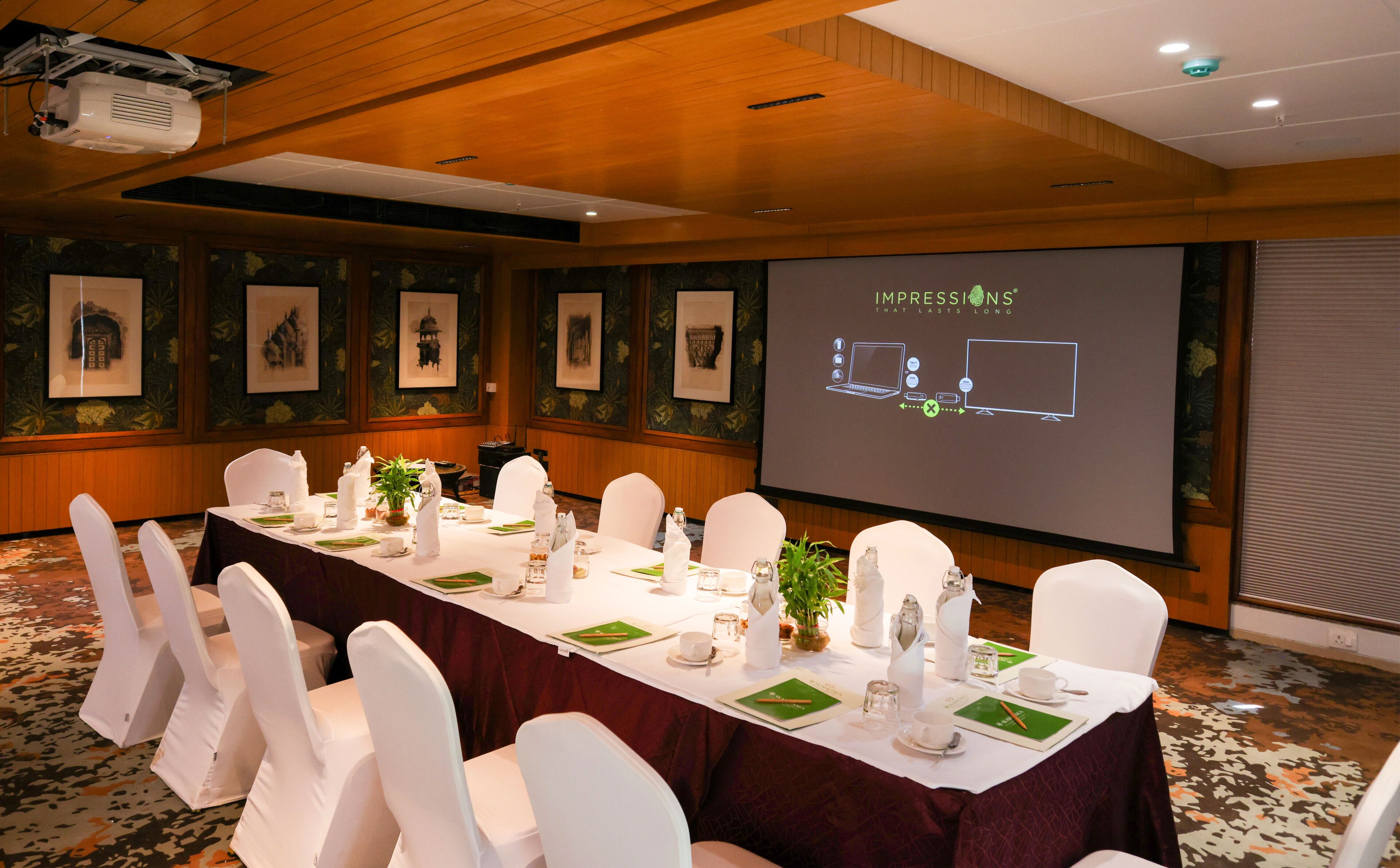 Un salón de conferencias con una larga mesa dispuesta para una reunión, sillas blancas, una pantalla de proyector y cuadros en las paredes.