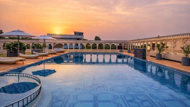 A swimming pool with a sunset view, surrounded by a covered walkway with umbrellas and a building with arches. - WelcomHeritage Akashganga Resort & Spa (Bichhiwara)